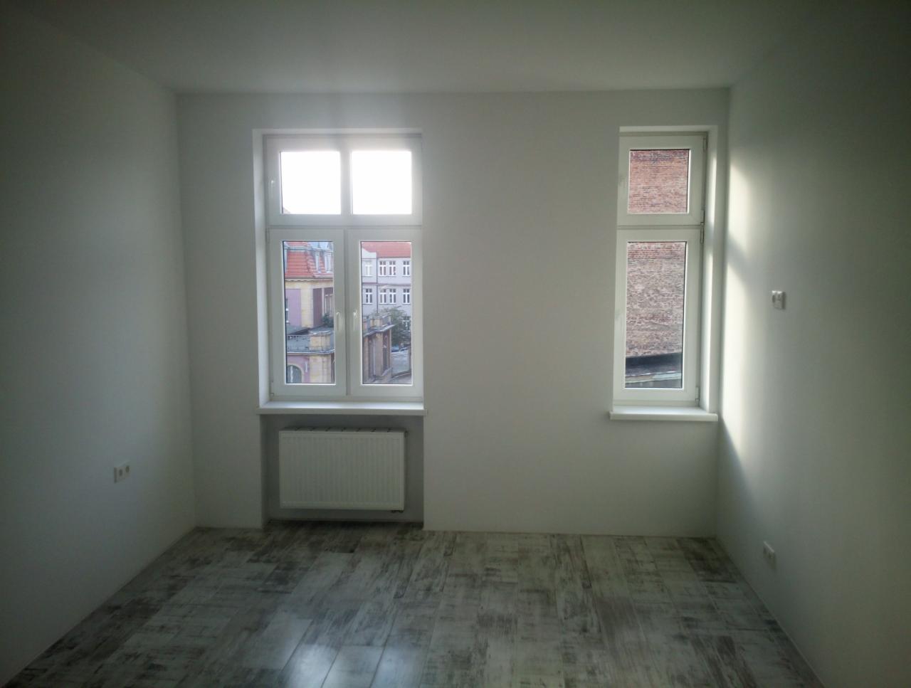 Pusty pokój z dwoma oknami o różnej wielkości, świeżo pomalowanymi ścianami na biało i szarą podłogą imitującą drewno, grzejnik pod większym oknem.
