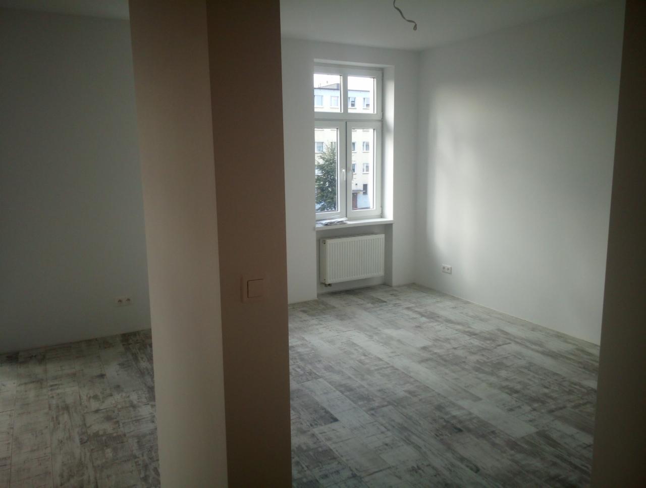 Pomalowane na biało wnętrze pokoju po remoncie z szarą podłogą imitującą panele, widoczne gniazdka elektryczne i grzejnik pod oknem.