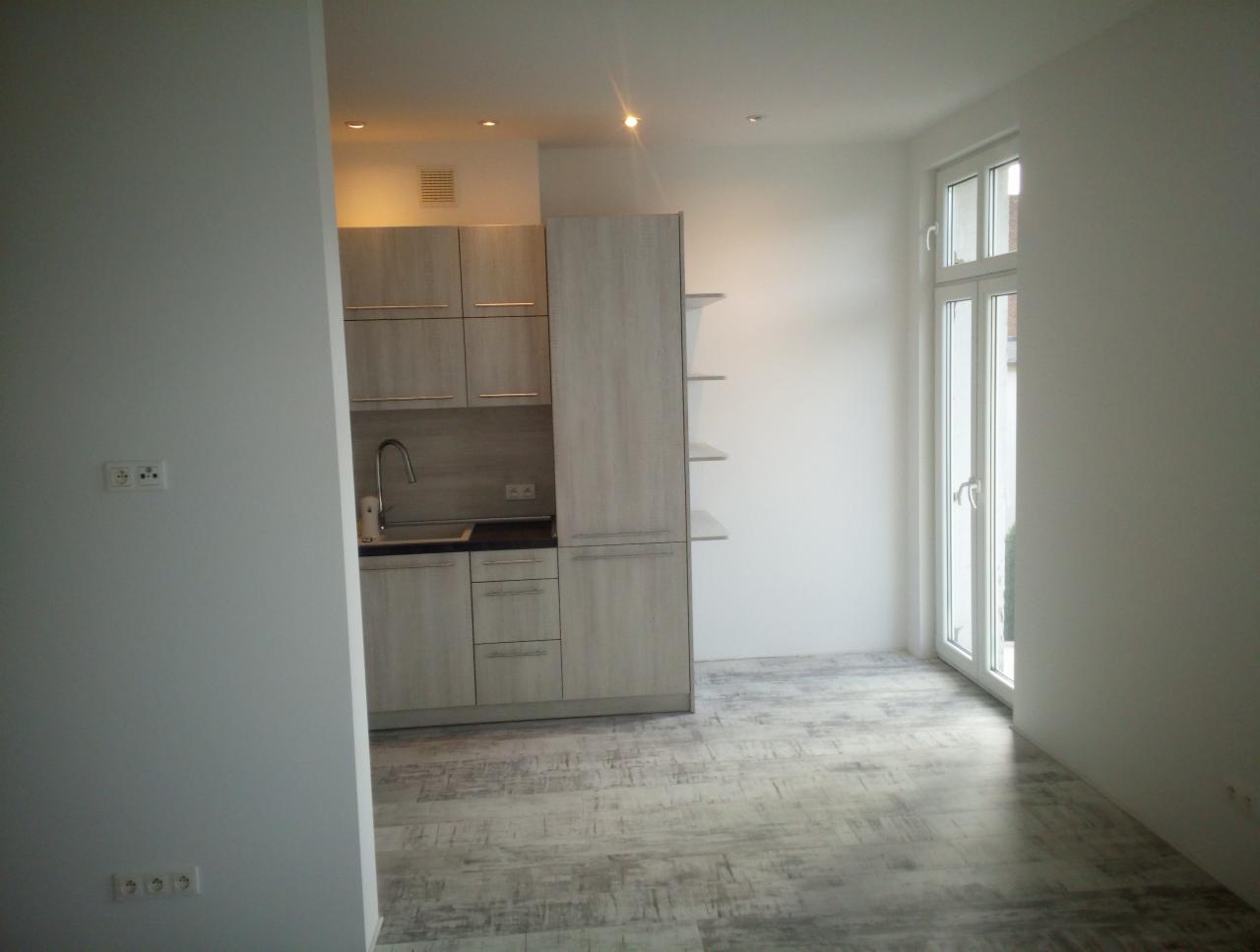 Jasna kuchnia z drewnianymi szafkami i szarymi panelami podłogowymi, widok na okno balkonowe, gniazdka elektryczne na ścianie.