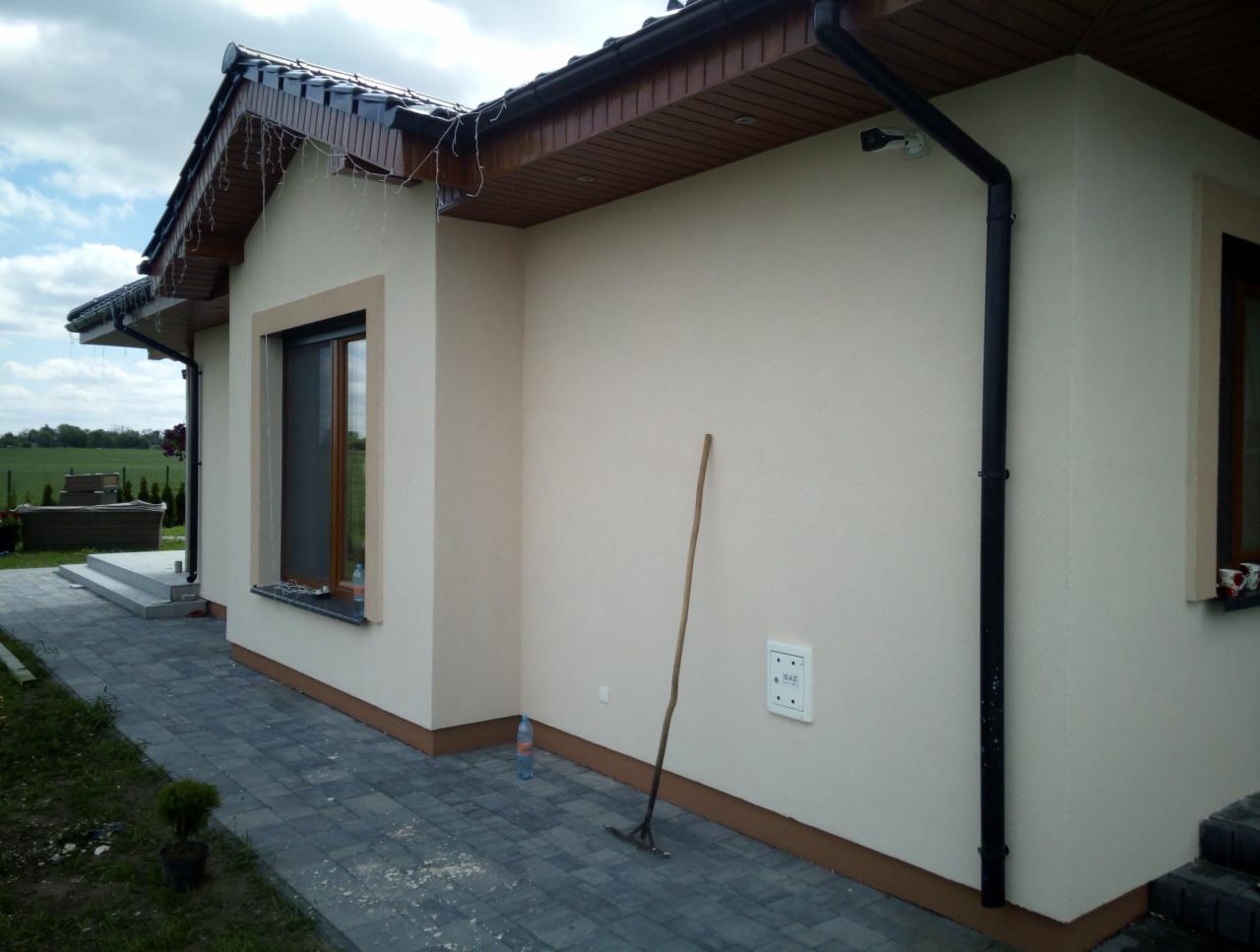 Wykończona elewacja domu jednorodzinnego w jasnym kolorze, widoczne okno z brązową ramą, czarna rynna, kostka brukowa na podjeździe i narzędzie oparty o ścianę.