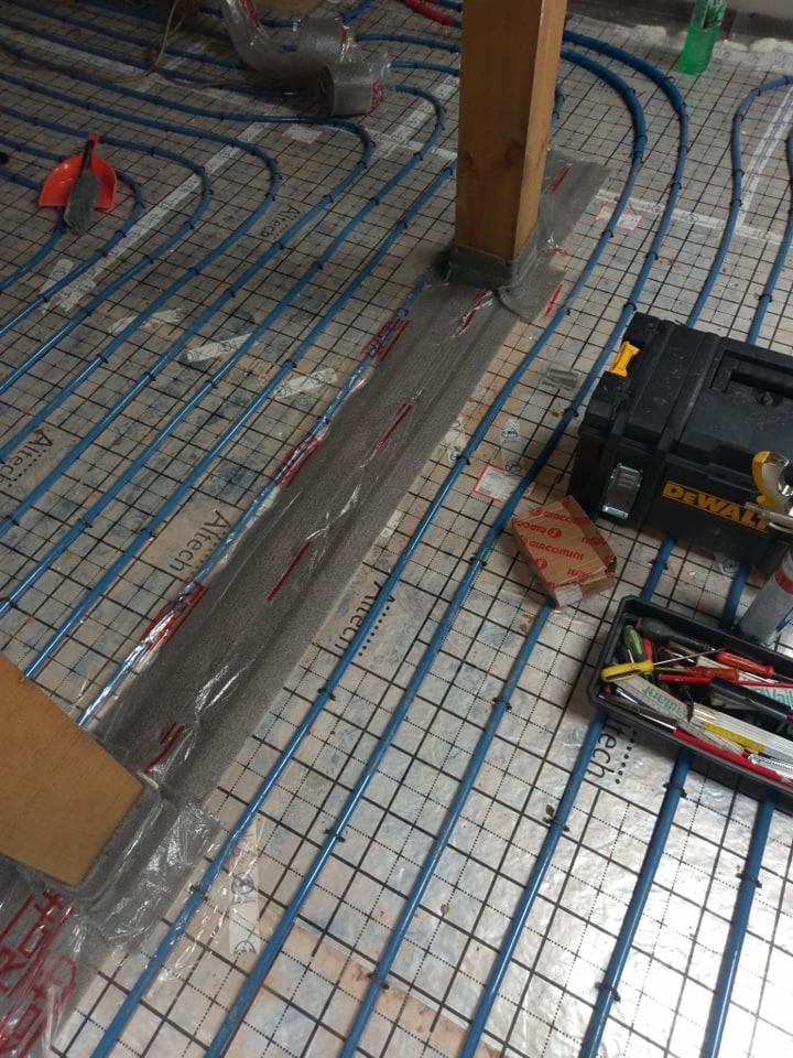 Montaż ogrzewania podłogowego: niebieskie rurki ułożone na siatce, narzędzia w skrzynce DeWalt, fragment izolacji wokół drewnianego słupa.