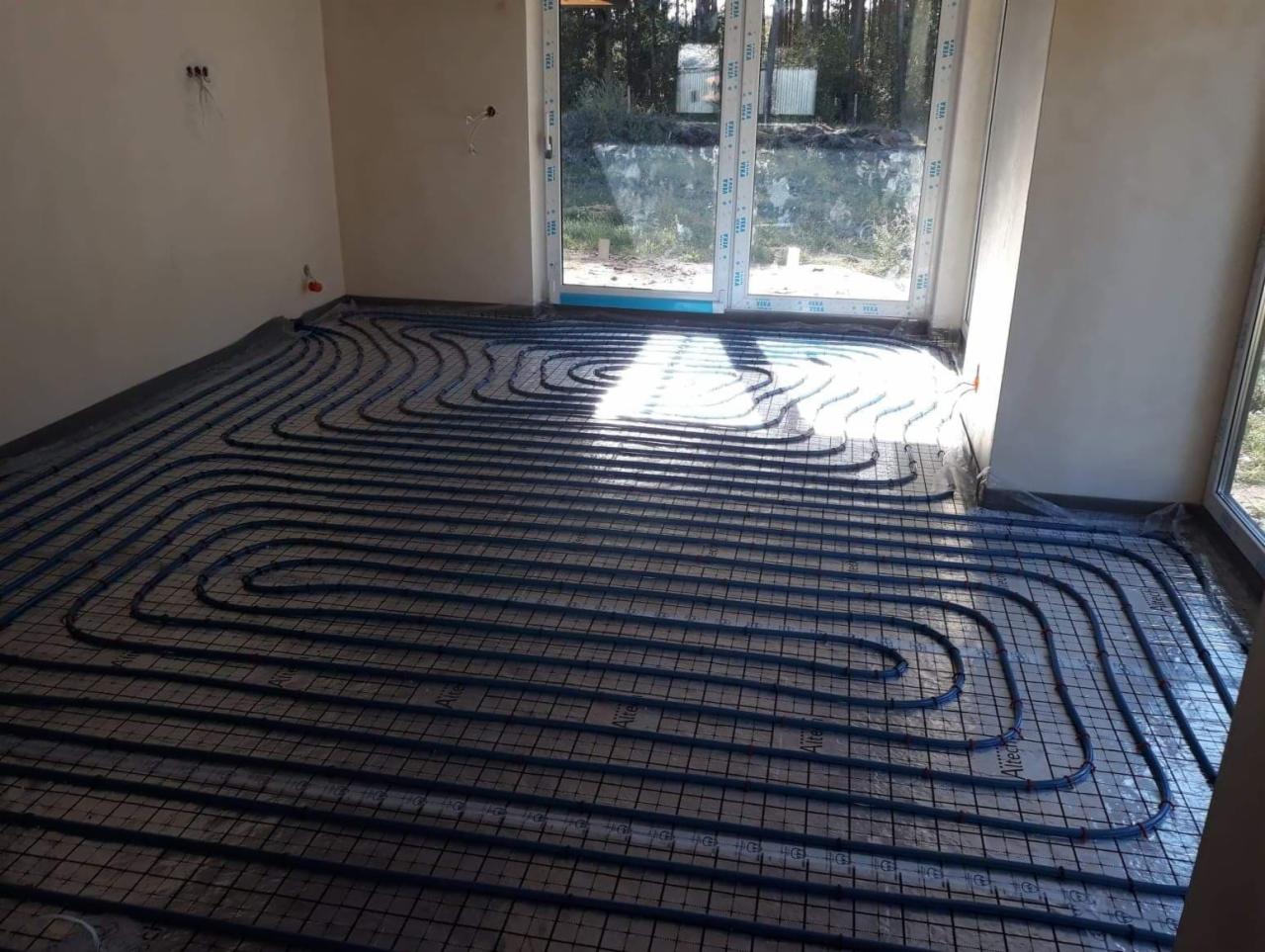 Instalacja ogrzewania podłogowego z rur w kolorze niebieskim na folii izolacyjnej w jasnym pomieszczeniu z dużym oknem tarasowym. Widoczne wystające przewody elektryczne na ścianie.