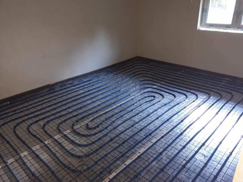 Rozłożone rury ogrzewania podłogowego na izolacji z siatką w jasnym pomieszczeniu, widoczne okno w rogu.