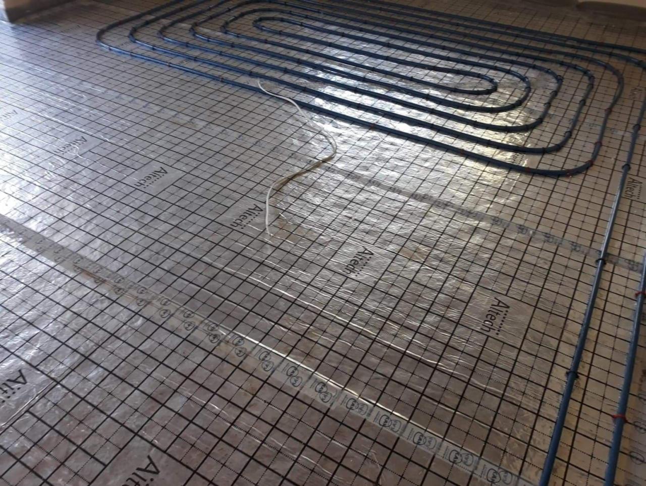 Instalacja ogrzewania podłogowego: rury grzewcze ułożone na folii z nadrukowaną siatką, widoczne mocowania rur.