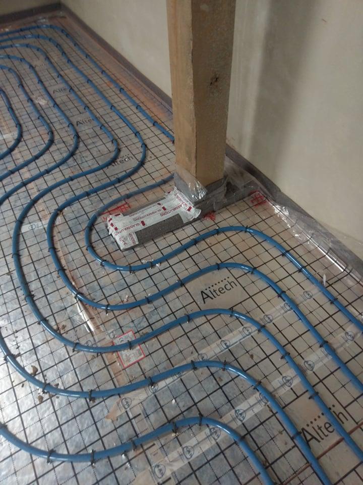 Niebieskie rury ogrzewania podłogowego ułożone na folii z nadrukowaną siatką, przymocowane czarnymi klipsami, widoczny fragment drewnianej kolumny zabezpieczonej folią, opakowanie styropianu...