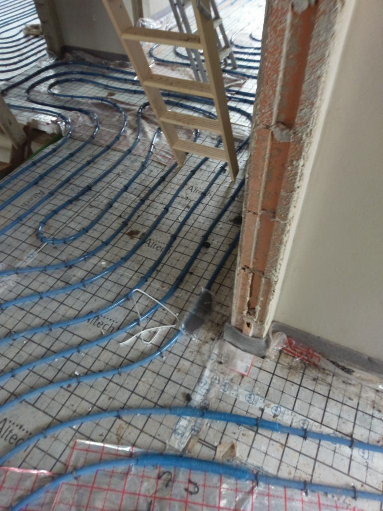 Montaż ogrzewania podłogowego: niebieskie rurki Altech ułożone na siatce, widoczna drabina i surowy filar w trakcie prac wykończeniowych.