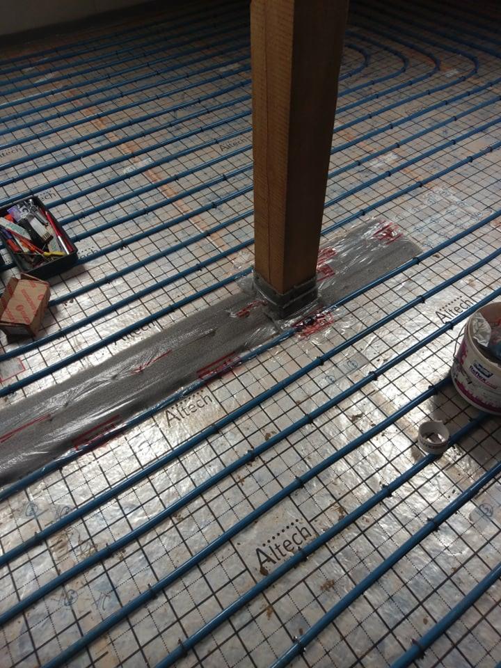 Montaż ogrzewania podłogowego: niebieskie rurki Altech ułożone na siatce, częściowo przykryte folią, z drewnianym słupem przechodzącym przez instalację.