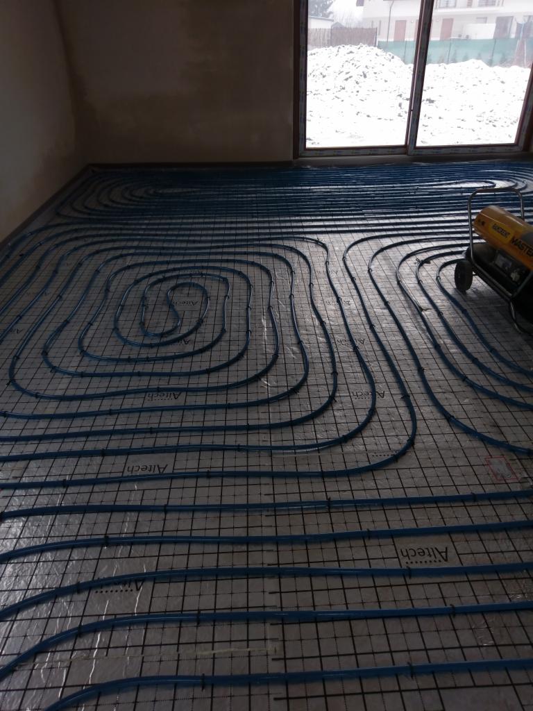 Niebieskie rury ogrzewania podłogowego ułożone spiralnie na siatce podłogowej w surowym wnętrzu, widoczny nagrzewnica powietrza i śnieg za oknem.