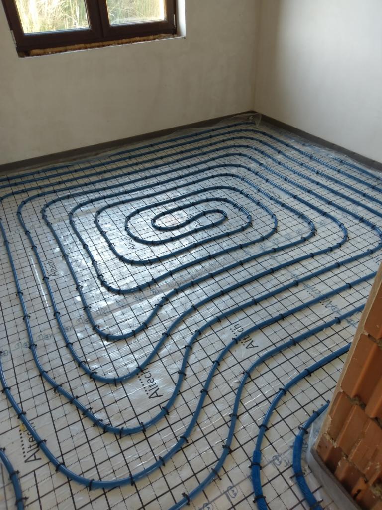 Niebieskie rury ogrzewania podłogowego ułożone spiralnie na siatce w surowym pomieszczeniu z widocznym oknem i fragmentem ceglanej ściany.