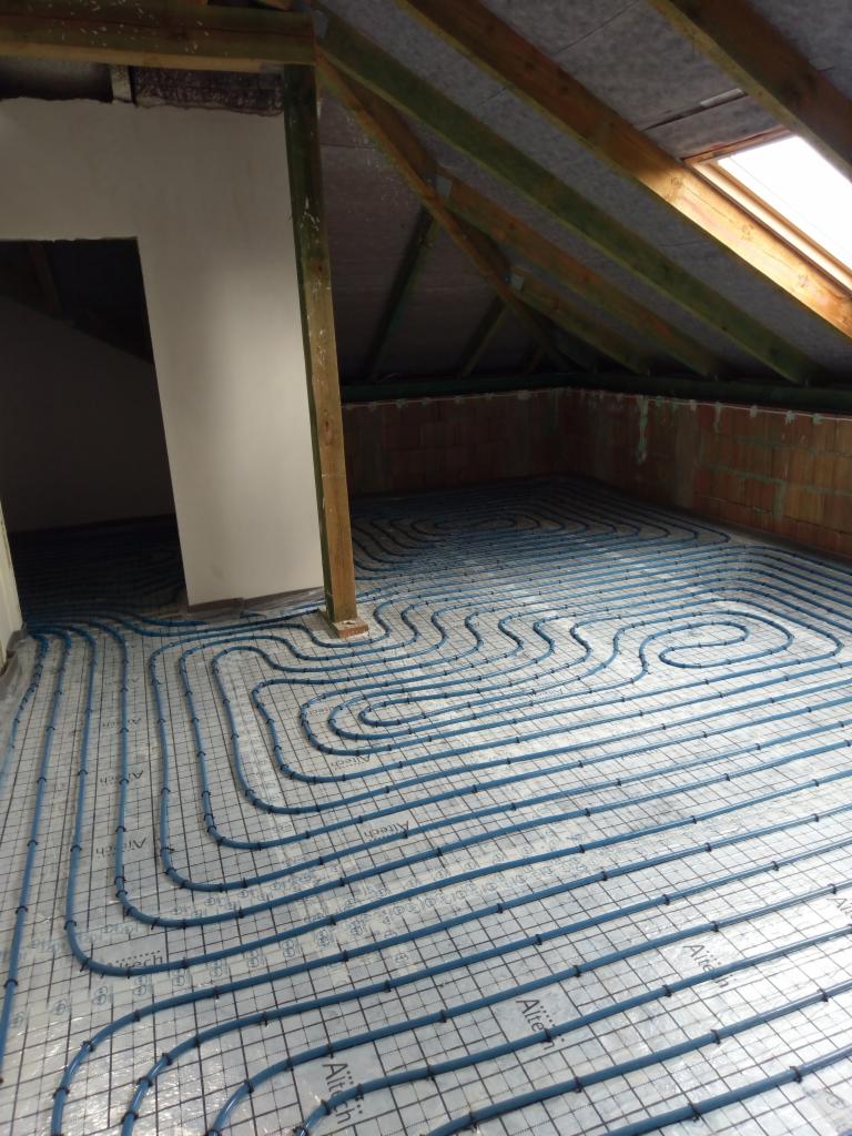Instalacja ogrzewania podłogowego z niebieskich rurek w surowym pomieszczeniu na poddaszu, widoczne krokwie i okno dachowe.