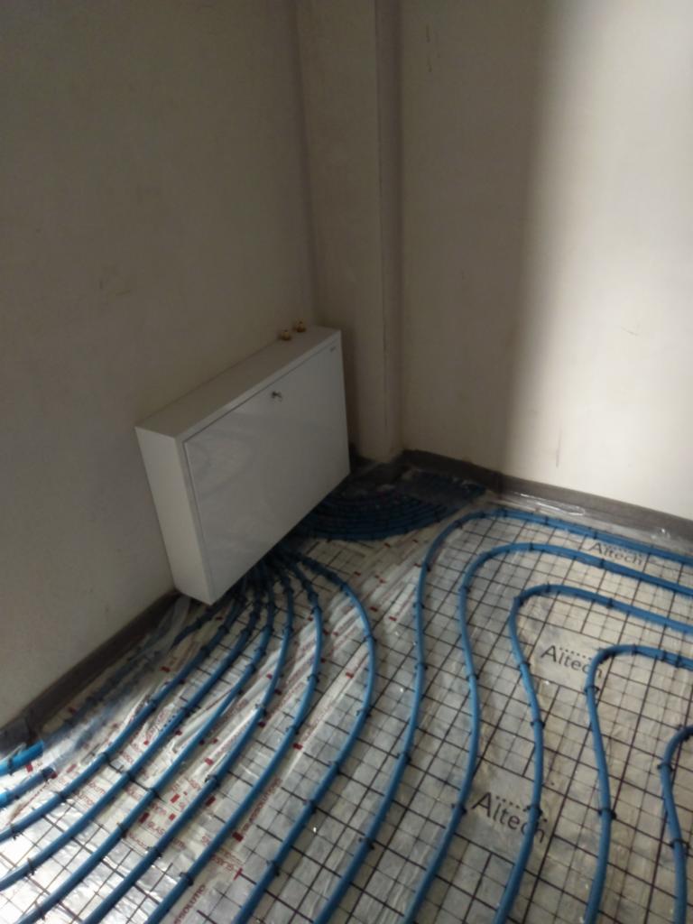 Rozłożona instalacja ogrzewania podłogowego z niebieskich rur na folii z siatką, obok biała szafka z zaworami na ścianie.