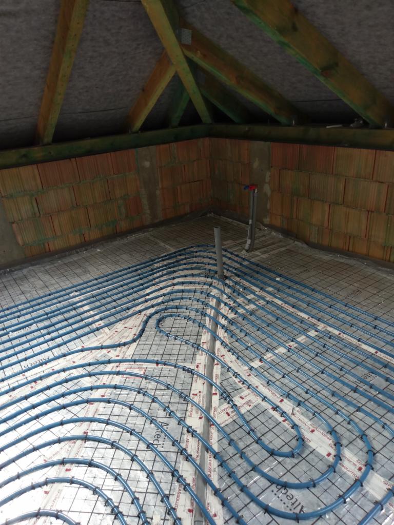Niebieskie rury ogrzewania podłogowego ułożone na izolacji z siatką w surowym stanie poddasza, widoczne ceglane ściany i drewniana konstrukcja dachu.