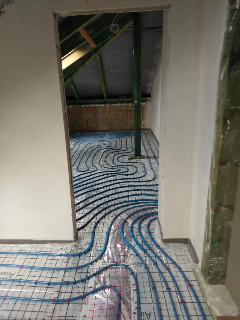 Instalacja ogrzewania podłogowego w surowym wnętrzu poddasza, widoczne nieotynkowane ściany i elementy konstrukcji dachu.