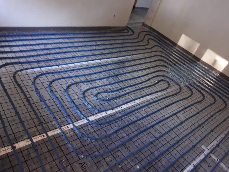 Rozłożona instalacja ogrzewania podłogowego z niebieskich rur na folii z nadrukiem siatki i logo Alitech, widok z góry na surową posadzkę w trakcie montażu.
