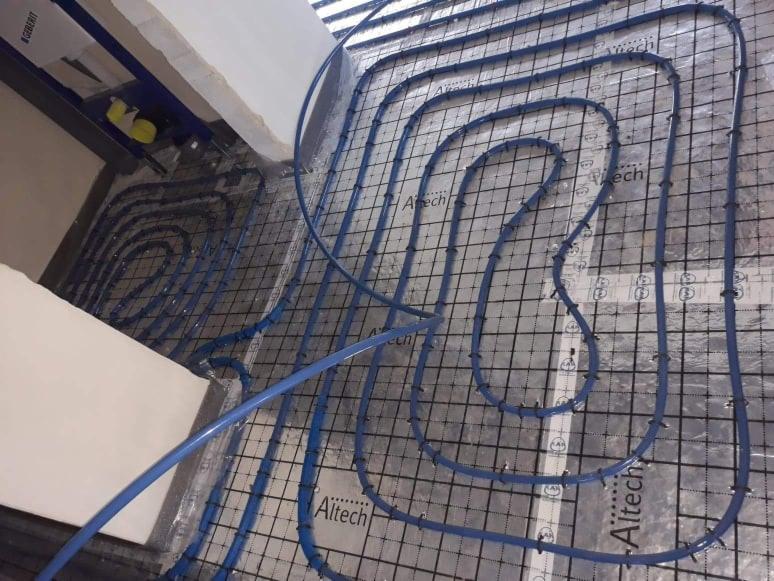 Niebieskie rury ogrzewania podłogowego ułożone na siatce Altech w trakcie montażu, widoczny fragment stelaża podtynkowego Geberit.