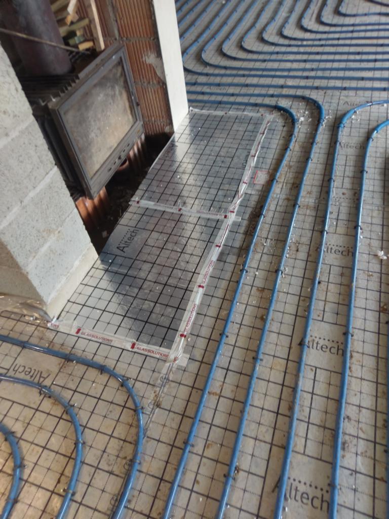 Montaż ogrzewania podłogowego: niebieskie rury ułożone na siatce z oznaczeniami Altech i Glassolutions, widoczny fragment kominka i surowa ściana.