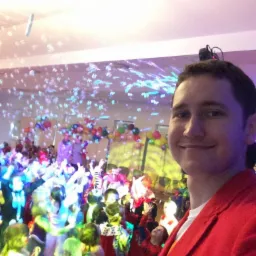 Selfie mężczyzny w czerwonej marynarce na tle bawiących się dzieci na imprezie z kolorowymi balonami i bańkami mydlanymi.