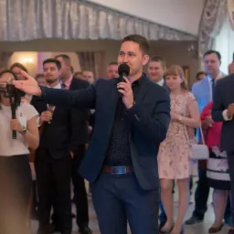 Mężczyzna w granatowym garniturze przemawia do zgromadzonych gości trzymając mikrofon, w tle widoczni ludzie z kieliszkami w dłoniach, rejestracja wydarzenia przez kamerzystę.