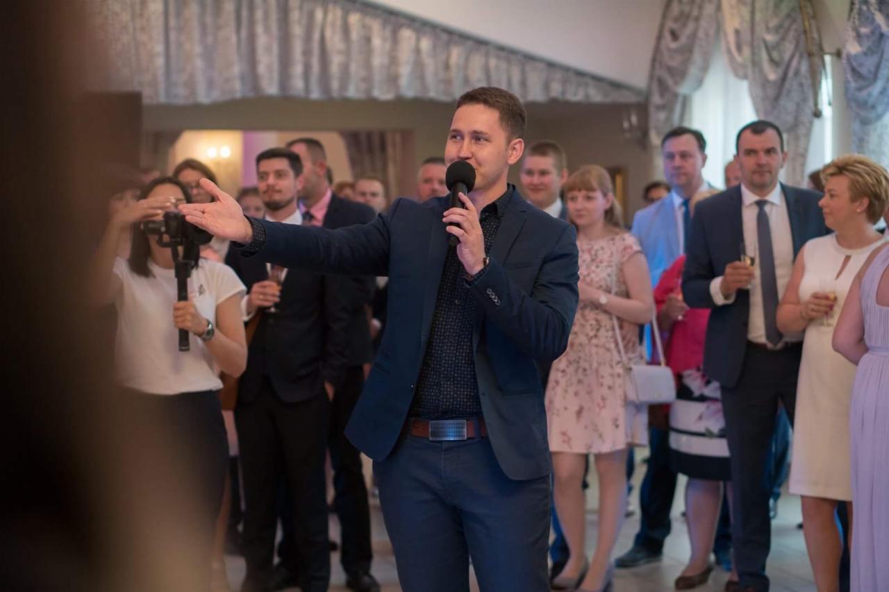 Mężczyzna w granatowym garniturze przemawia do zgromadzonych gości trzymając mikrofon, w tle widoczni ludzie z kieliszkami w dłoniach, rejestracja wydarzenia przez kamerzystę.