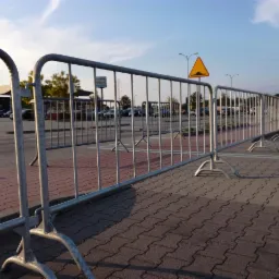 Długi rząd metalowych barierek ochronnych z poprzecznymi prętami, ustawionych na kostce brukowej parkingu w słoneczny dzień, z żółtym znakiem ostrzegawczym o nierówności drogi w tle.