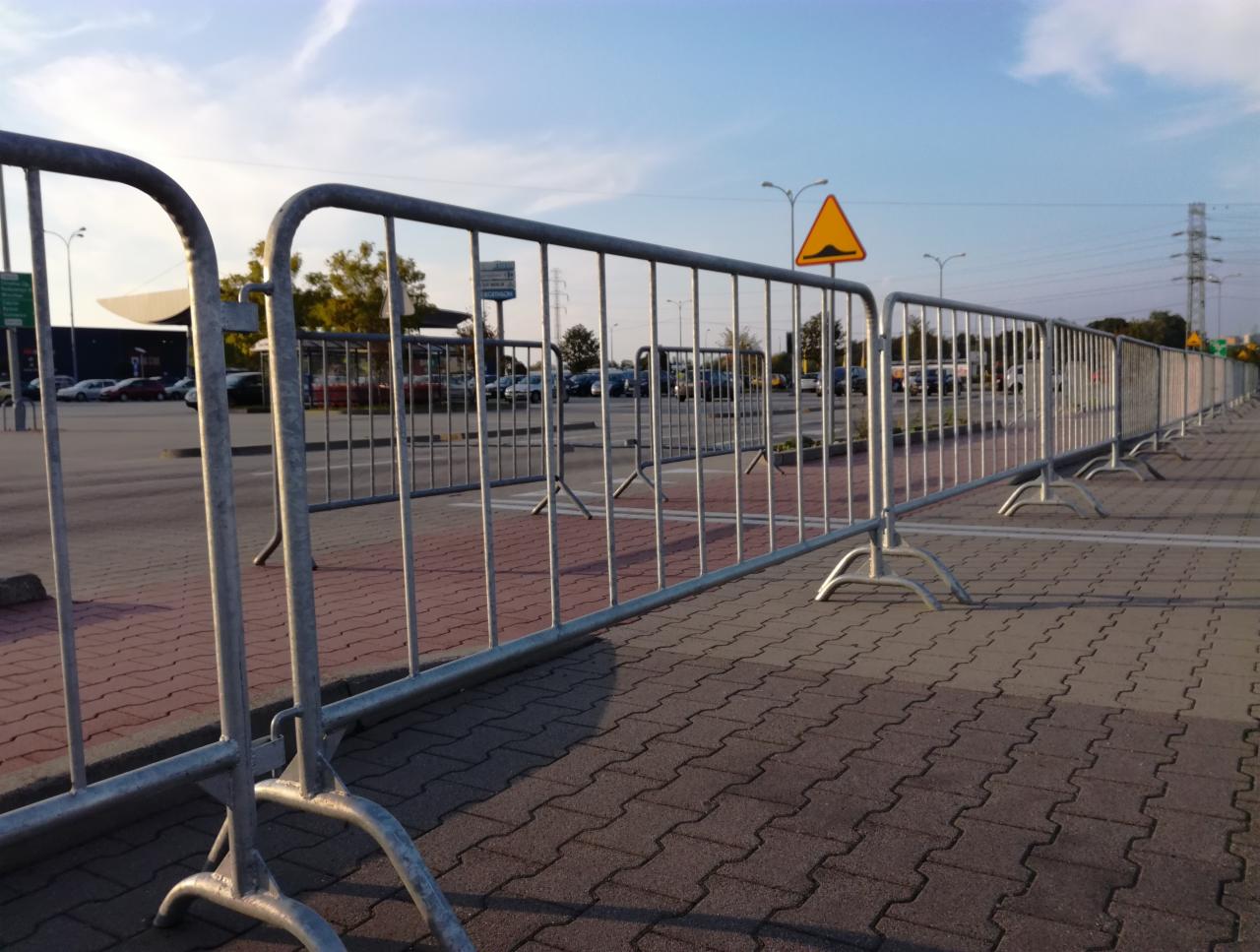 Długi rząd metalowych barierek ochronnych z poprzecznymi prętami, ustawionych na kostce brukowej parkingu w słoneczny dzień, z żółtym znakiem ostrzegawczym o nierówności drogi w tle.