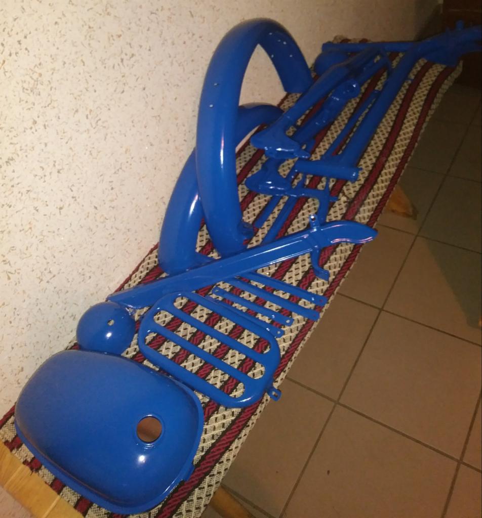 Elementy motocykla, w tym błotnik, rama i zbiornik, świeżo pomalowane na niebiesko, ułożone na wzorzystej ławce.