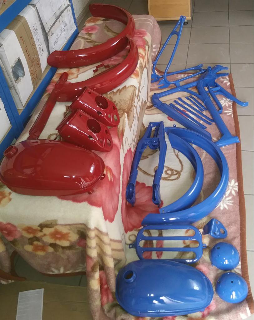 Części motocyklowe po renowacji: czerwone błotniki, zbiornik paliwa i obudowy lamp oraz niebieska rama, widelec i inne elementy, ułożone na kwiecistej narzucie.