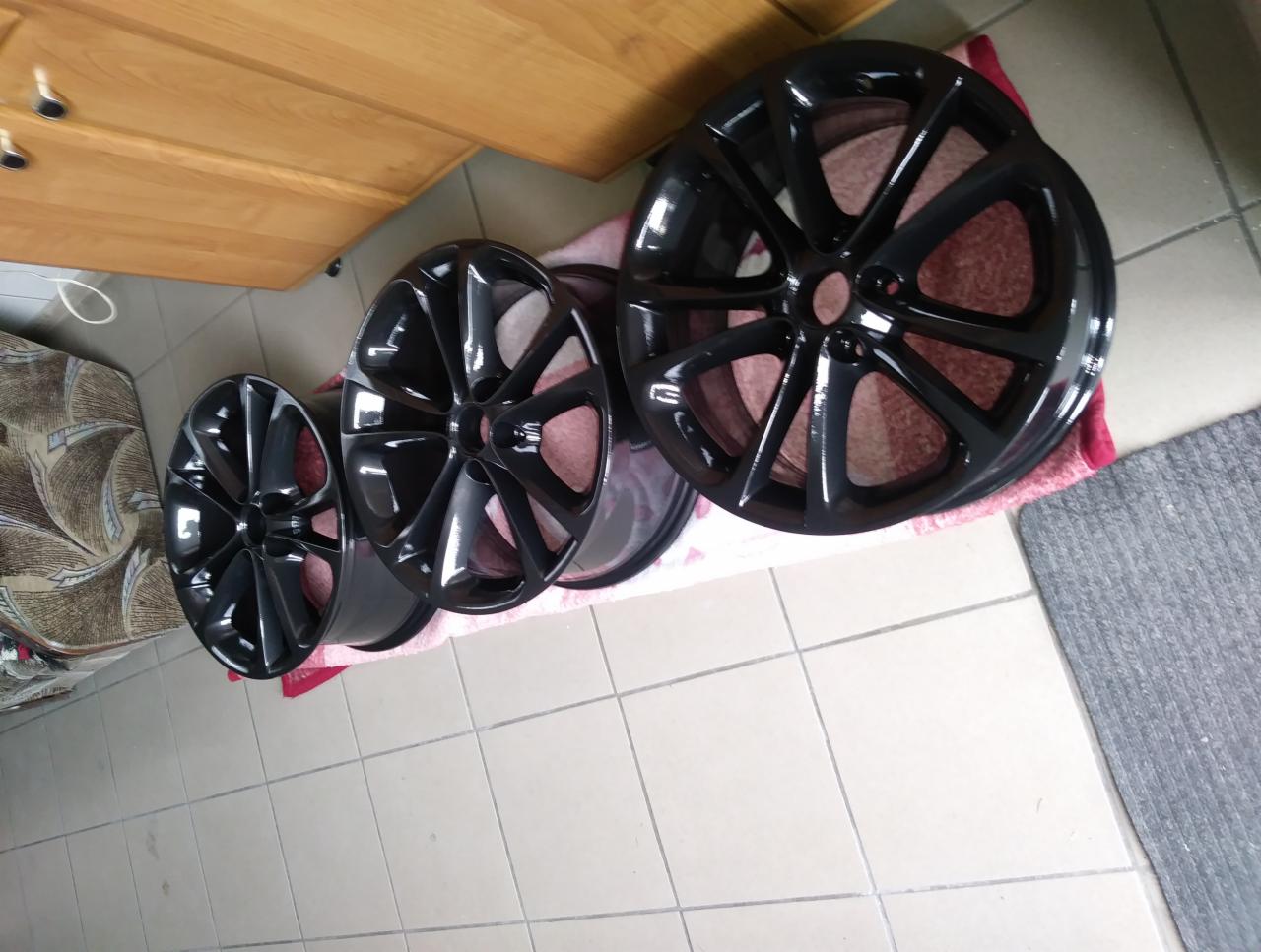 Cztery czarne, błyszczące felgi aluminiowe ułożone rzędami na podłodze, na tle beżowych płytek i fragmentu szafki.