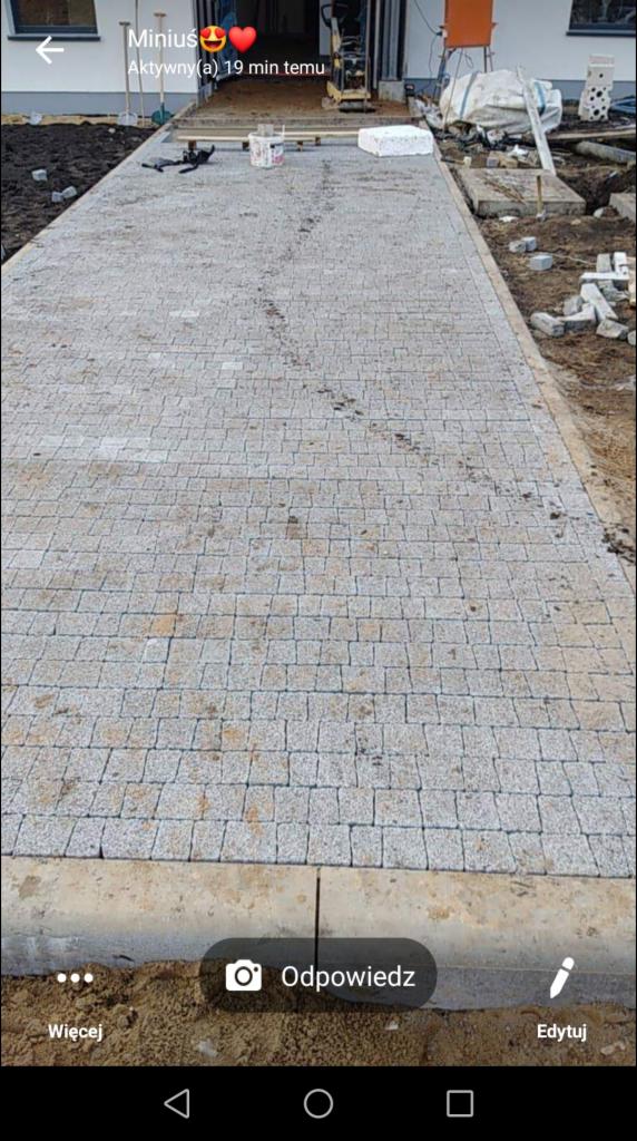 Świeżo ułożona ścieżka z jasnoszarej kostki granitowej, widoczna ubijarka w tle, obrzeża betonowe, teren jeszcze nieuporządkowany.