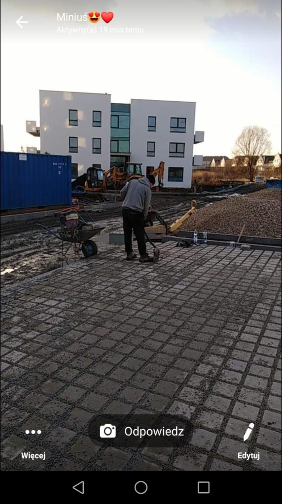 Pracownik zagęszcza wibratorem ułożoną kostkę brukową na posesji, w tle budynek mieszkalny i maszyny budowlane.