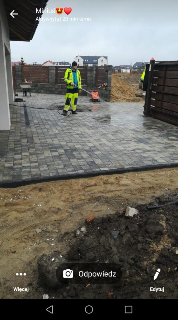 Pracownik w odblaskowym kombinezonie czyści nowo ułożoną szarą kostkę brukową za pomocą myjki ciśnieniowej. Widoczne świeże fugi, przygotowany teren pod dalsze prace i ogrodzenie posesji.