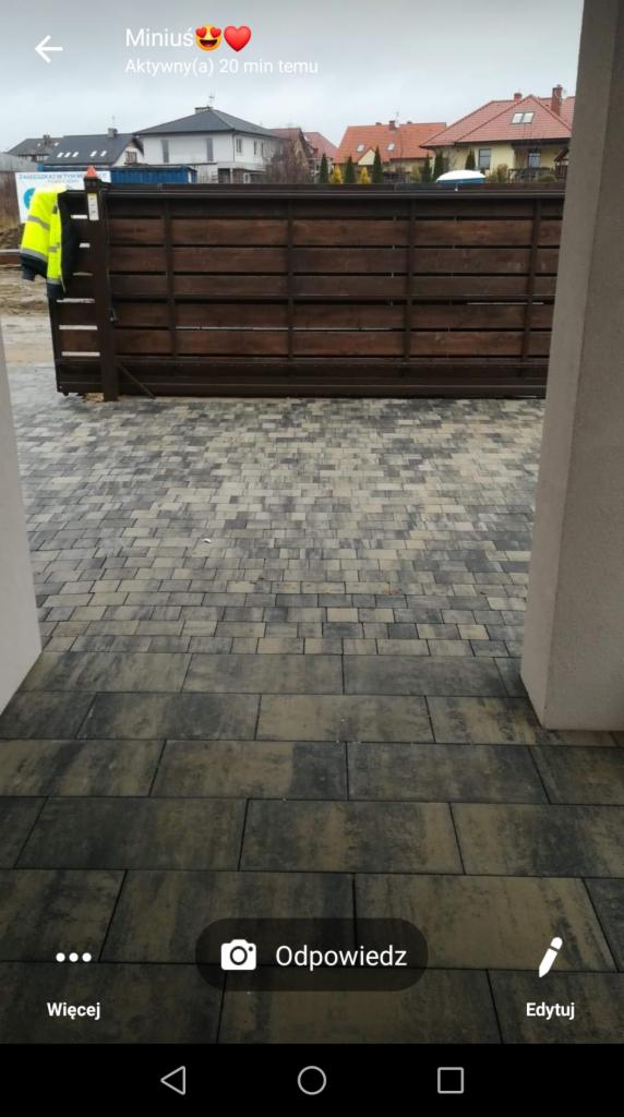 Ułożona kostka brukowa w odcieniach szarości i beżu prowadząca do bramy przesuwnej z drewnianymi panelami, widok z wnętrza budynku.