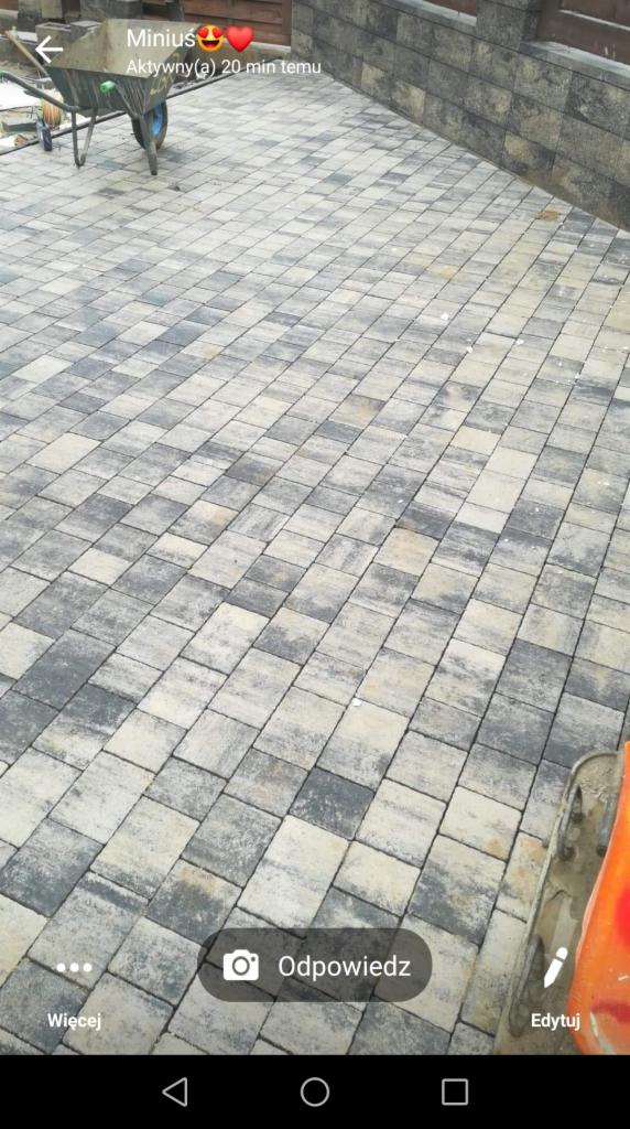 Ułożona kostka brukowa w odcieniach szarości i beżu, z widocznym w tle fragmentem muru z kostki oraz taczką. Widoczne ślady użytkowania.