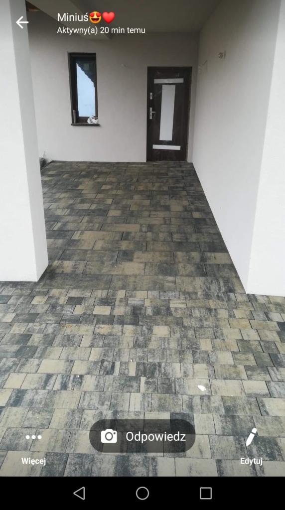 Ułożona kostka brukowa w odcieniach szarości i beżu tworząca wzór na podjeździe przed budynkiem z białymi ścianami i ciemnymi drzwiami.