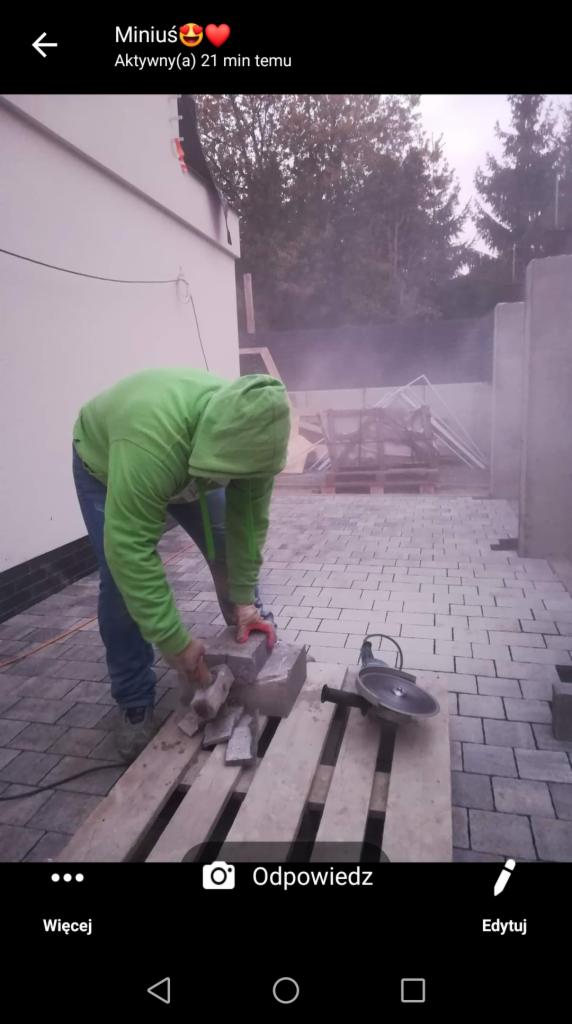 Pracownik w zielonej bluzie układa kostkę brukową na posesji, widoczna paleta z kostką i szlifierka kątowa, w tle budynek i drzewa.