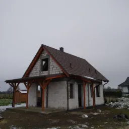 Dom w trakcie budowy, z brązowym dachem i drewnianą konstrukcją werandy, widoczne bloczki betonowe, częściowo ośnieżony teren.