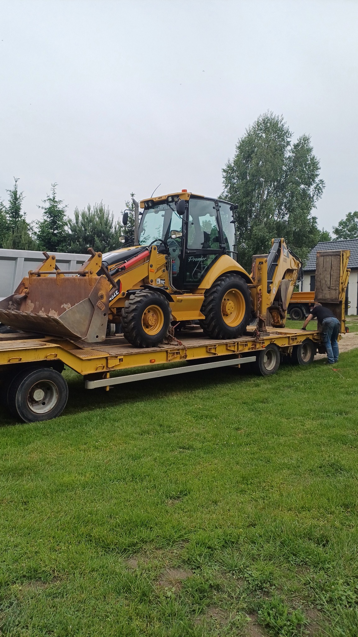 Żółta koparko-ładowarka CAT 432E na niskopodwoziowej przyczepie, zabezpieczana przez mężczyznę w jeansach. Trawa i budynki w tle, pochmurne niebo.