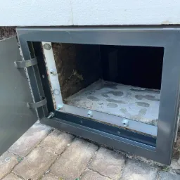 Szara metalowa obudowa okienka piwnicznego z otwartymi drzwiczkami, widoczne mocowania śrubowe i fragment ściany z kamienną teksturą oraz kostka brukowa na zewnątrz.