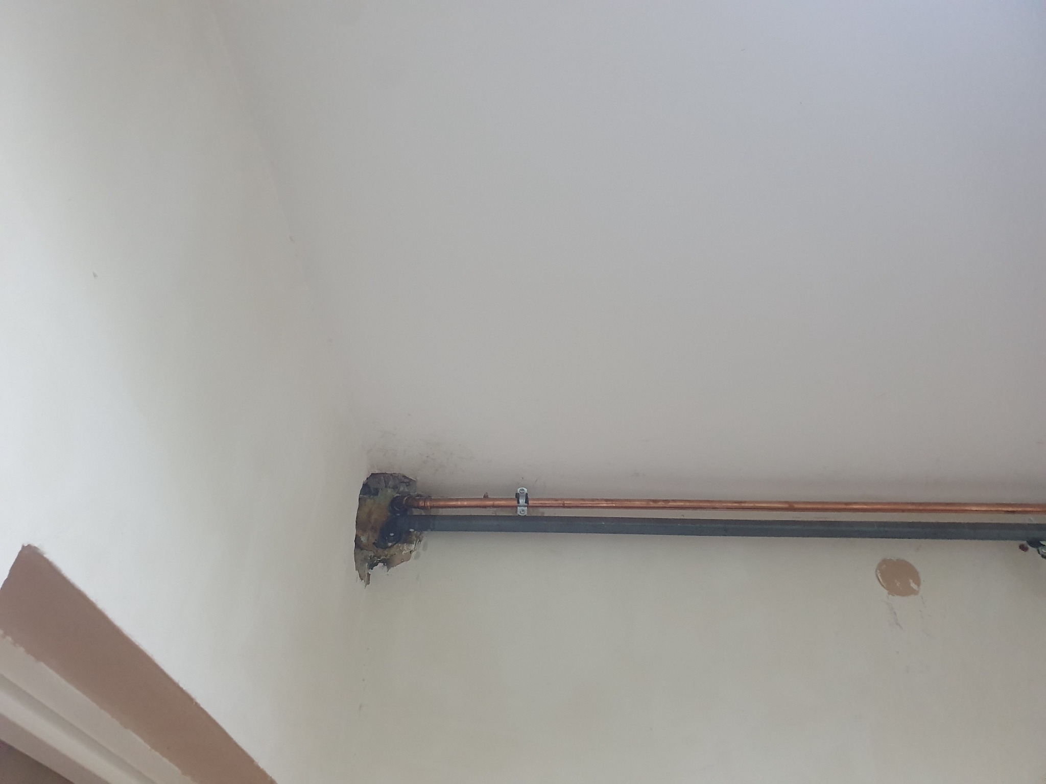 Miedziana rura grzewcza przymocowana do ściany z widocznym uszkodzeniem tynku w miejscu przejścia przez ścianę, dodatkowo widoczna szara rura instalacyjna.