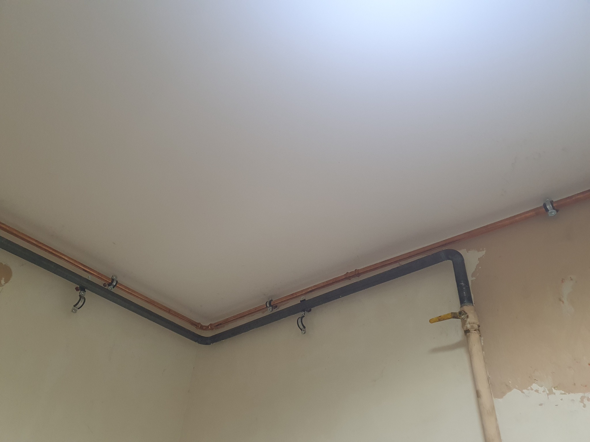 Instalacja gazowa i miedziana pod sufitem, widoczne rury w kolorze miedzi i czarnym, mocowane do ściany za pomocą uchwytów, z zaworem gazowym z żółtą rączką.