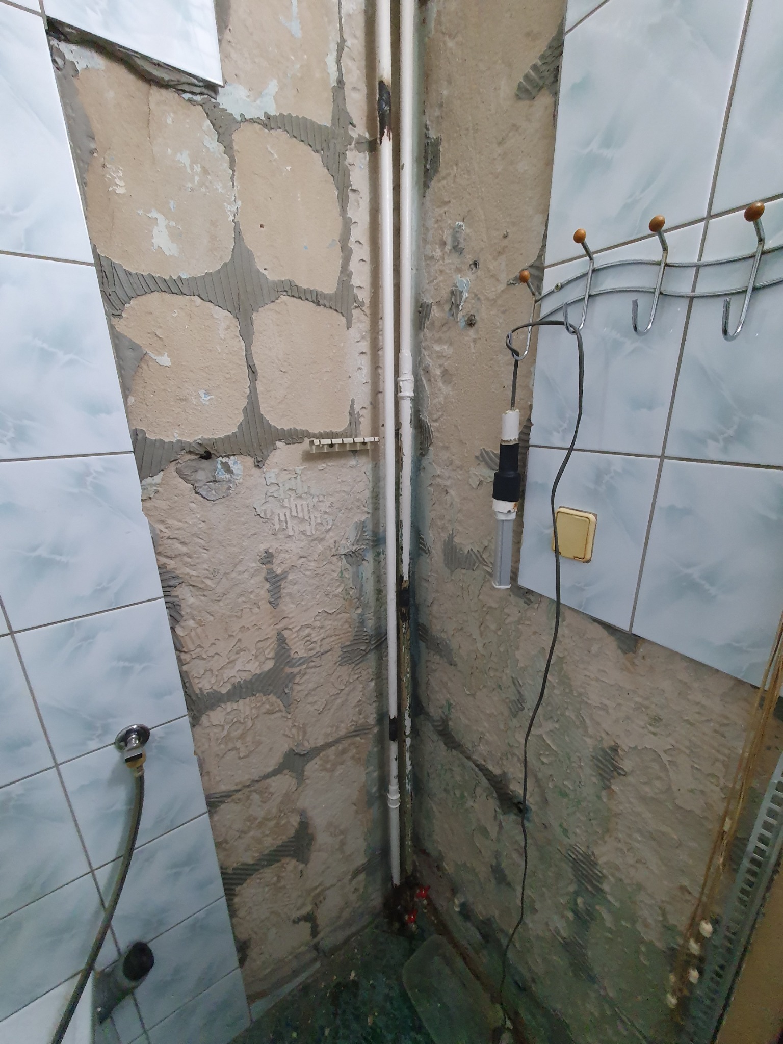 Fragment łazienki w trakcie remontu: częściowo położone jasne płytki, odsłonięte fragmenty ściany z resztkami starej zaprawy, rury instalacyjne i wiszący haczyk na ręczniki.