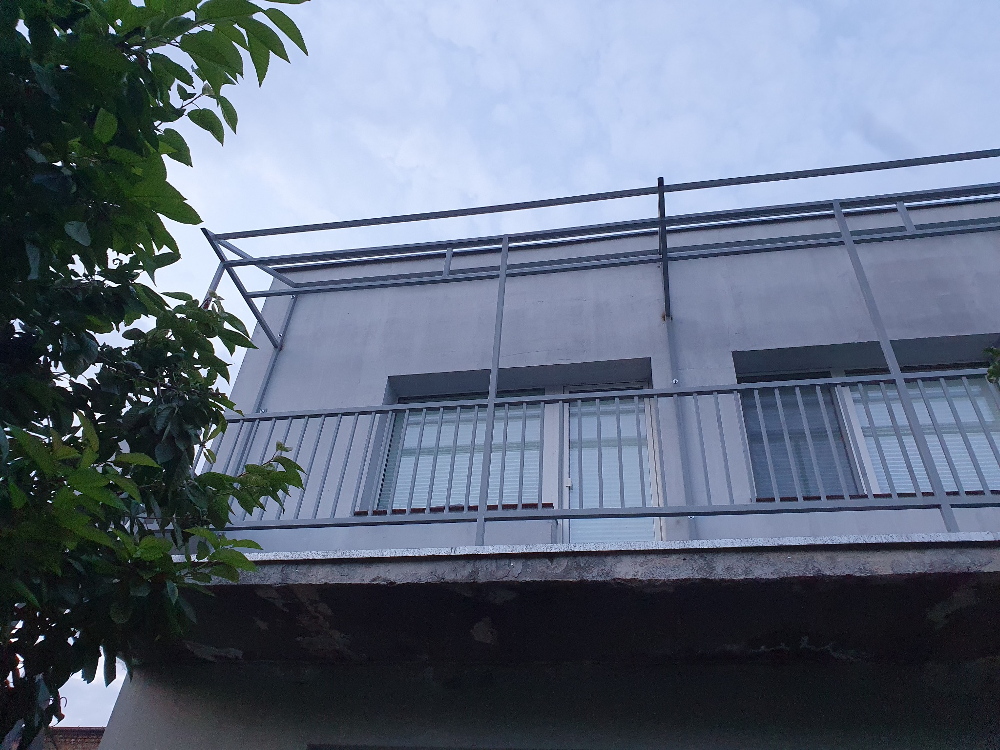 Szary balkon z metalową balustradą i zadaszeniem, widok z dołu na tle pochmurnego nieba, częściowo zasłonięty przez gałęzie drzewa po lewej stronie.