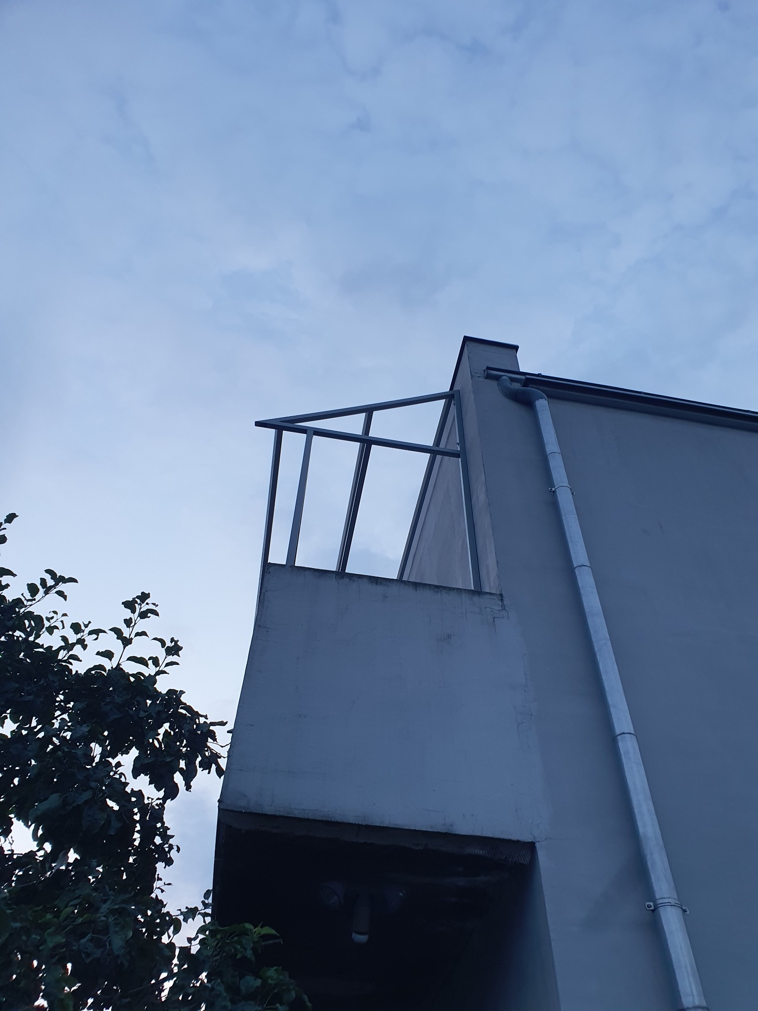 Niski kąt ujęcia budynku z metalową konstrukcją balkonu w surowym stanie, widoczna rynna i fragment drzewa na tle pochmurnego nieba.