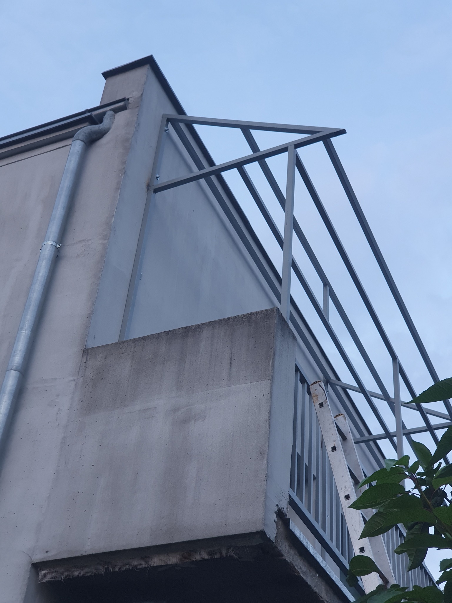 Metalowa konstrukcja balkonu z widoczną rynną i fragmentem drabiny opartą o balustradę, ujęcie z dołu.