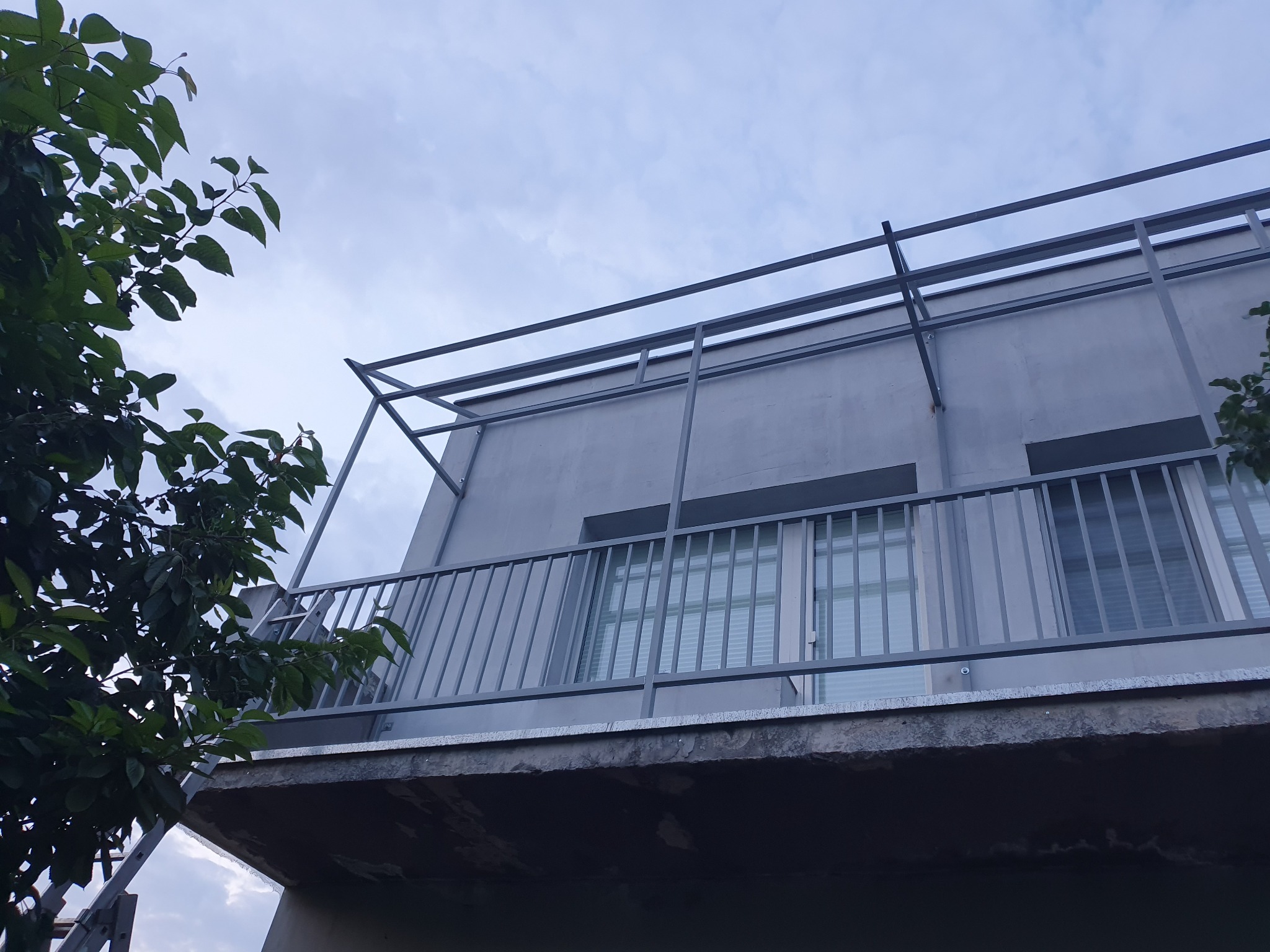 Metalowa konstrukcja balkonu z szarymi barierkami, widok z dołu na tle pochmurnego nieba i fragmentu drzewa.