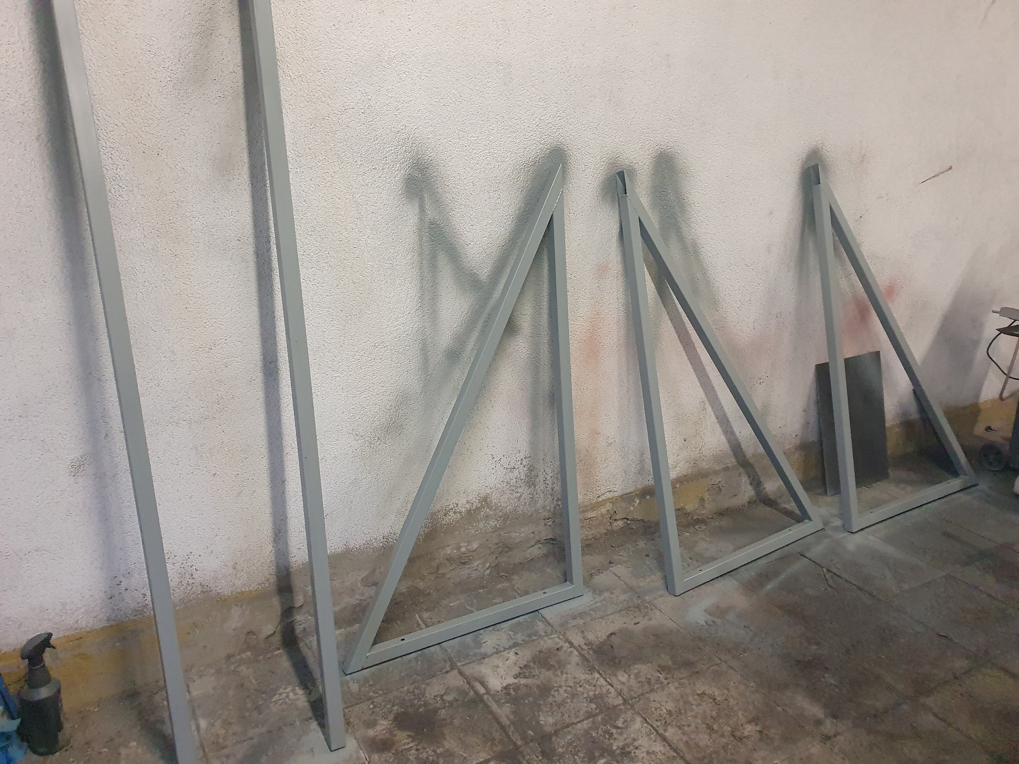 Szare, metalowe konstrukcje trójkątne o profilu zamkniętym, ustawione rzędem przy ścianie o chropowatej fakturze, prawdopodobnie po malowaniu.
