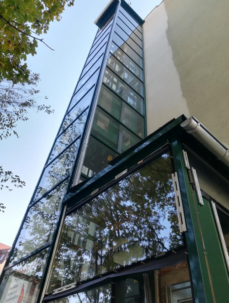 Zewnętrzna, przeszklona konstrukcja windy przy budynku, z widocznymi elementami metalowymi i odbiciem drzew w szybach. Ujęcie z dołu, podkreślające wysokość konstrukcji.