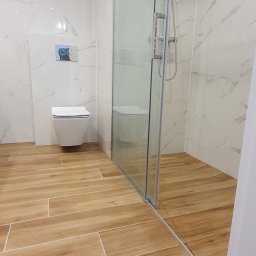 Adam Grygo - Nowoczesna łazienka z kabiną prysznicową typu walk-in, podwieszaną toaletą i drewnopodobną podłogą z płytek, wykończona marmurowymi płytami na ścianach.