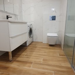 Adam Grygo - Nowoczesna łazienka z białymi marmurkowymi płytkami ściennymi, drewnopodobną podłogą, wiszącą toaletą z chromowanym przyciskiem spłukiwania, białą szafką umywalkową na nóżkach i pralką w rogu.