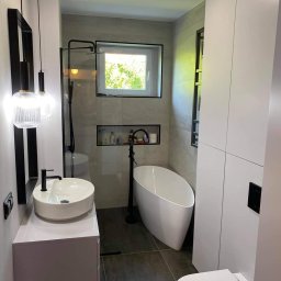 Adam Grygo - Nowoczesna łazienka z wolnostojącą wanną, kabiną prysznicową ze szklaną ścianką, okrągłą umywalką nablatową z czarną armaturą i wiszącą toaletą, oświetlona wiszącymi lampami z żarówek dekoracyjnych.